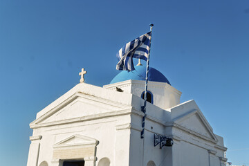 church of Agios Nikolaos with blue dome located on a stone pier with blue benches and a Greek flag capturing the iconic Cycladic architecture of Mykonos.