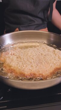 Close-up of breaded pork cutlet frying in hot oil in a stainless steel pan in a home kitchen, vertical format