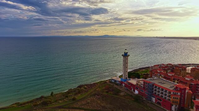Golden rays illuminate the coastline as a lighthouse oversees gentle waves, surrounded by charming buildings. The scenery captivates with its serene beauty and warm evening glow.