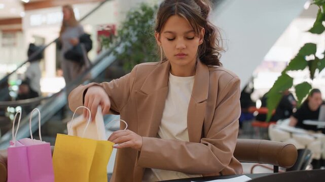 shopper carefully sorting her paper purchase bags, young customer reviewing receipts and items near escalator, retail shopper methodically inspecting her colorful bags and receipts after shopping