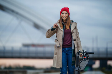 Woman in a beige coat and red hat smiles while standing beside her scooter on an overcast day near...