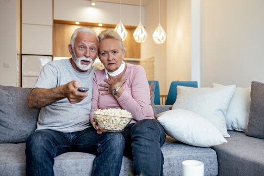 Elderly couple watching a thrilling movie at home and enjoying popcorn on a cozy sofa in the evening