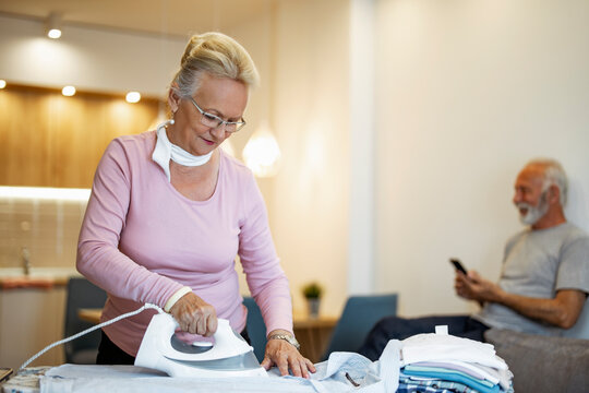 Senior woman ironing clothes in a cozy living room while an older man relaxes on the couch and uses his phone
