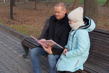 Father and daughter look at photo album on a park bench in autumn.