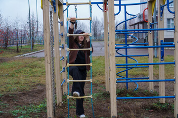 Pensive teenager in a brown jacket climbing a playground rope ladder on a gloomy autumn day.