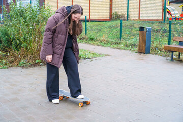 Young woman in a brown jacket learning to ride a skateboard in a park on a cool day.
