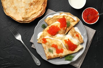 Tasty crepes with red caviar and sour cream on black textured table, flat lay