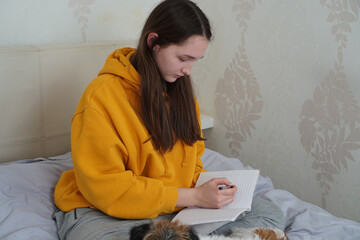 Teenage girl in yellow hoodie studying and writing in a notebook on her bed at home.