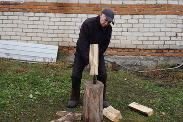 Senior man in a cap splitting firewood on a chopping block in the backyard