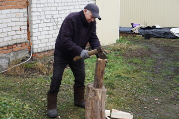 Elderly man in warm clothes chopping firewood with an axe on a stump in a rustic backyard.