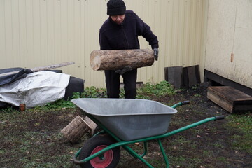 Man in warm clothes loading a heavy log into a wheelbarrow in a backyard.