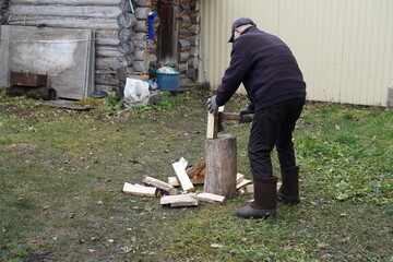 Senior man chopping firewood with an axe in a rural backyard next to a log cabin.