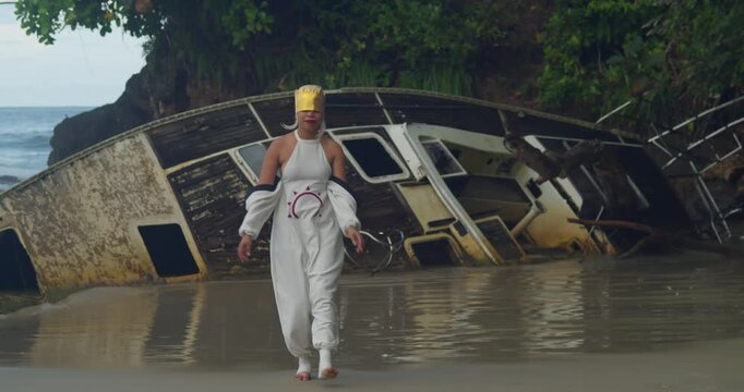 A wide shot of a girl in costume walking on the wet sand, her reflection visible in the water under a moody Caribbean sky.