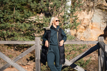 Blonde woman wearing sunglasses and a black vest standing on a wooden bridge, enjoying the autumn nature of Latvia.