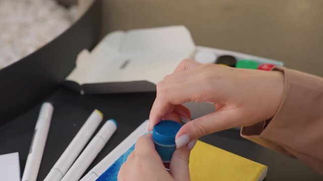 lady twisting open small blue paint container, woman carefully opens her blue paint jar on table, an individual gently unscrews blue paint container amidst scattered art supplies on tabletop