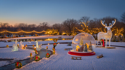Enchanting winter holiday light display in a snowy park with a large snow globe and illuminated reindeer at dusk.