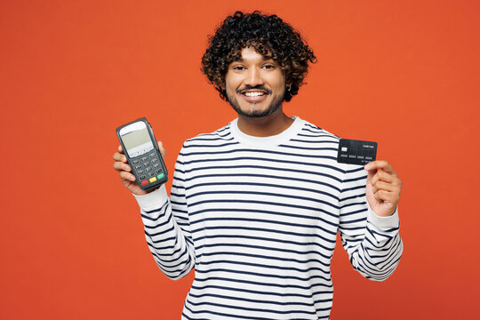 Young happy Indian man he wear striped sweatshirt casual clothes hold wireless modern bank payment terminal to process acquire credit card isolated on plain red orange background. Lifestyle concept. - Powered by Adobe
