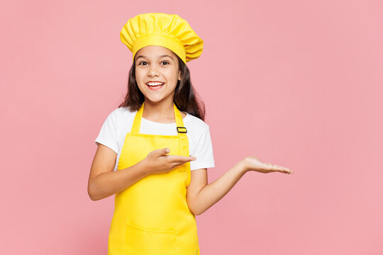 Small kid child girl housewife chef cook baker mother's helper wear yellow apron toque cap point index finger aside on blank area mock up isolated on pastel plain pink background Cooking food concept