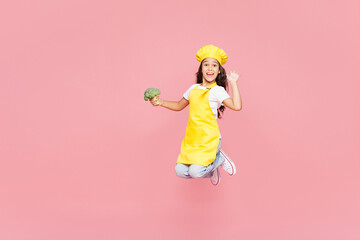Full body side view small kid child girl housewife chef cook baker mother's helper wear yellow apron toque cap jump high hold in hand broccoli isolated on plain pink background. Cooking food concept.
