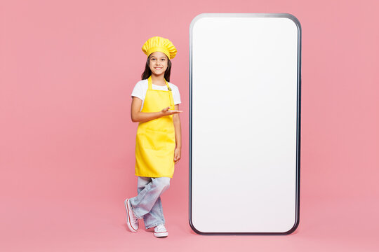 Full body small kid child girl housewife chef cook baker mother helper wear yellow apron toque cap big huge blank screen area mobile cell phone isolated on plain pink background. Cooking food concept.