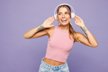 Young happy woman wears pink tank top casual clothes listen to music in headphones look aside on blank empty area isolated on plain pastel light purple background studio portrait. Lifestyle concept.