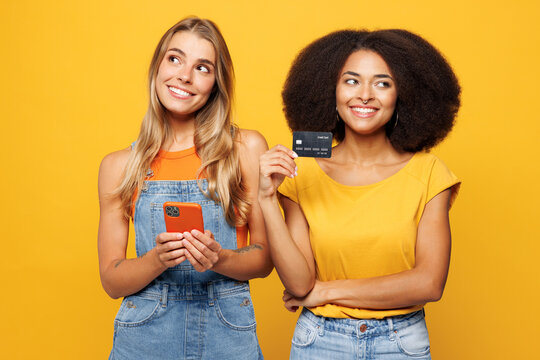 Two young friends buddies women wear denim casual clothes together using mobile cell phone hold credit bank card shopping online order delivery booking tour isolated on plain yellow orange background.