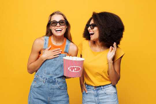Two young smiling happy friends buddies women in denim casual clothes 3d glasses watch movie film together hold bucket of popcorn in cinema isolated on plain yellow orange background studio portrait