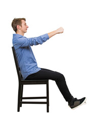 Side view full length portrait of confident teenage guy driver, pretending to drive an imaginary transportation, while sitting on a chair with arms stretched as holding the steering wheel
