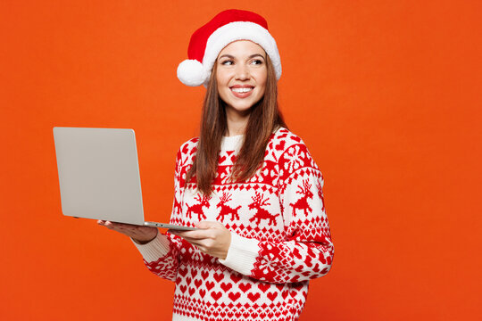 Merry young IT woman wear red knitted Christmas sweater Santa hat posing hold use work on laptop pc computer look aside isolated on plain orange background. Happy New Year celebration holiday concept.