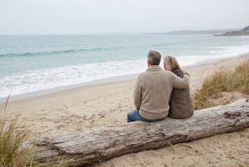 Mature Couple Sitting on Driftwood Log Watching Ocean Waves in Winter