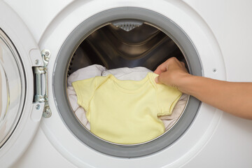 Young adult mother hands holding and unloading yellow baby bodysuit from opened white washing machine. Closeup. Newborn clothes care. Front view.