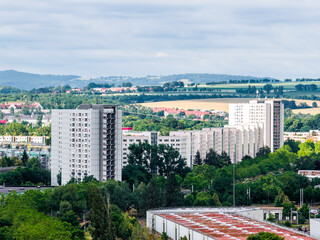 View of a residential area with panel buildings set against rolling hills, mixed with industrial zones, dense greenery, and a broad rural panorama.