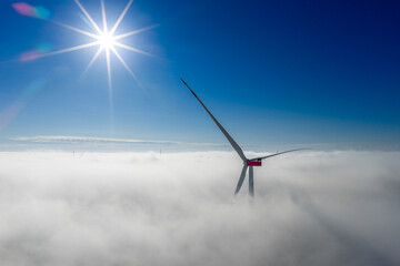 A wind turbine rises through a dense cloud layer, with the bright sun shining above the clear sky. The turbine stands majestically above the sea of fog.