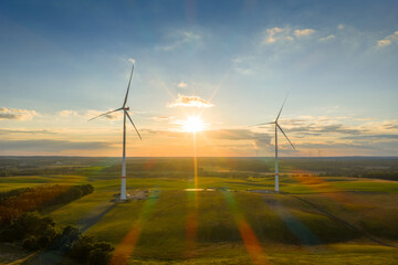 Two wind turbines stand in a wide landscape at sunset. The low sun illuminates the fields and hills, creating warm, colorful light reflections.