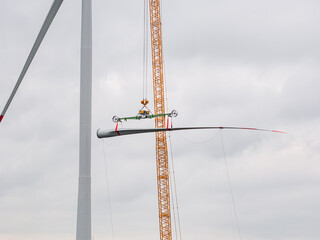 A rotor blade is lifted by a crane for installation on a wind turbine. The large structure is assembled step by step under a gray sky.
