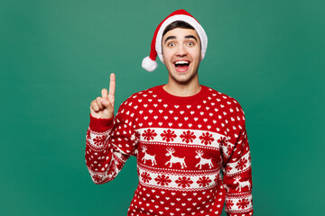 Merry young man wear red warm cozy Christmas sweater Santa Claus hat posing holding index finger up...