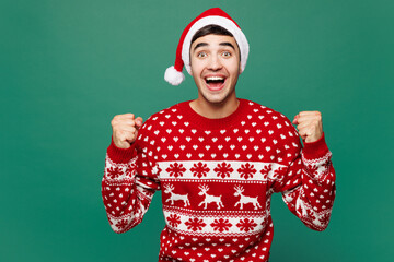 Merry young man wear red warm cozy Christmas sweater Santa Claus hat posing do winner gesture...