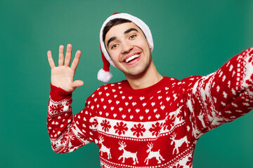 Merry young man wears red Christmas sweater Santa Claus hat posing oing selfie shot pov on mobile cell phone waving hand isolated on plain green background. Happy New Year celebration holiday concept.