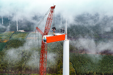 A large crane installs a wind turbine in dense fog. Technicians work high above the landscape while more turbines fade into the misty background.
