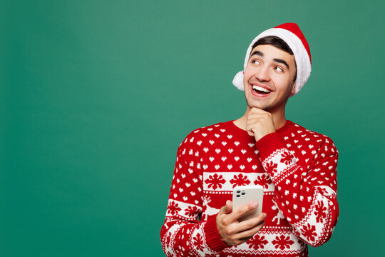 Merry young minded man wears red warm cozy Christmas sweater Santa Claus hat posing hold in hand use mobile cell phone isolated on plain green background. Happy New Year celebration holiday concept.