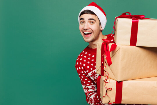 Merry young surprised man wear red warm cozy Christmas sweater Santa Claus hat posing hold stack present box with gift ribbon bow isolated on plain green background Happy New Year celebration concept