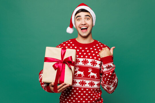 Merry young man in red Christmas sweater Santa Claus hat posing hold present box with gift ribbon bow point finger aside isolated on plain green background Happy New Year celebration holiday concept - Powered by Adobe