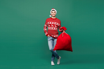 Full body merry young smiling man wearing red warm cozy Christmas sweater hat posing hold Santa...