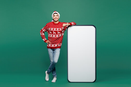 Full body merry young man wear red Christmas sweater Santa Claus hat posing next to big huge blank screen mobile cell phone area smartphone isolated on plain green background. Happy New Year concept.