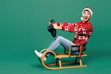 Full body merry young fun man wear red warm cozy Christmas sweater Santa Claus hat posing ride sled...