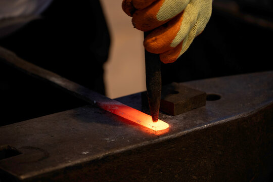 Doha, Qatar - December 01, 2025: Close up of a blacksmith shaping a glowing hot metal piece on an anvil, using a chisel while wearing protective gloves. - Powered by Adobe