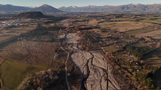 Aerial over the braided, dry riverbed of Wanaka River at sunset, surrounded by brown rugged Southern Alps.