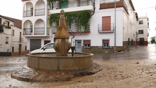 Water and mud fill a street during a severe flood in a Spanish village. A white car struggles to navigate past an ornamental stone fountain.