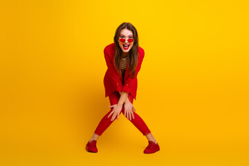 Adorable woman in stylish red suit and sunglass posing playfully against vibrant yellow background