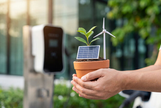 Hands holding potted plant with mini solar panel and wind turbine near electric car charger outdoors, clean energy and sustainable technology concept for eco friendly transportation and green living